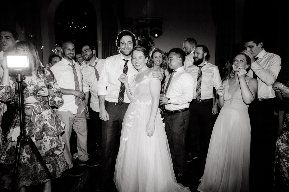 The bride and the groom are standing close to each other, smiling, while  many guests are around them.