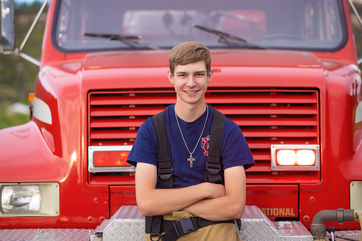 high school senior boy wearing his bunker gear sitting on the front of a fire truck with the lights on
