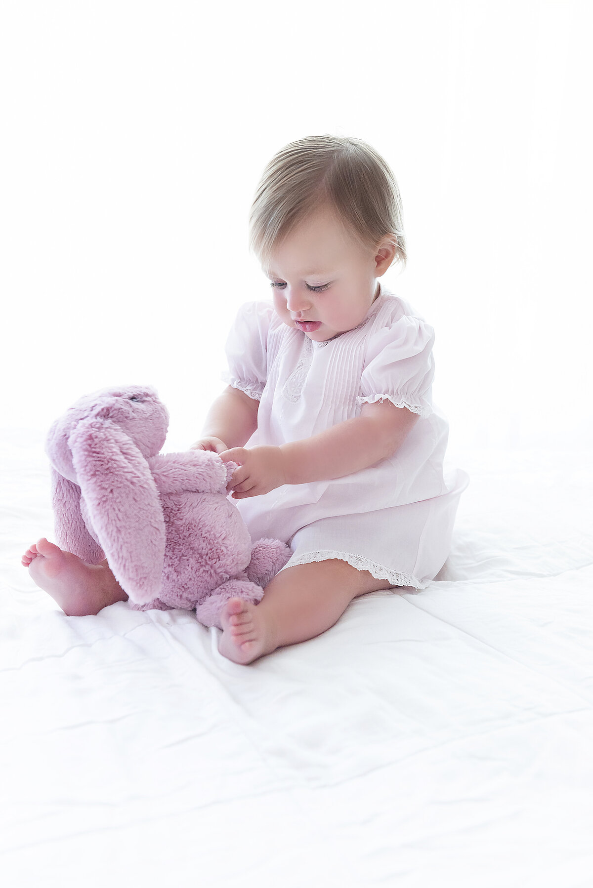 A toddler girl in a white dress sits on a bed playing with a pink stuffed bunny