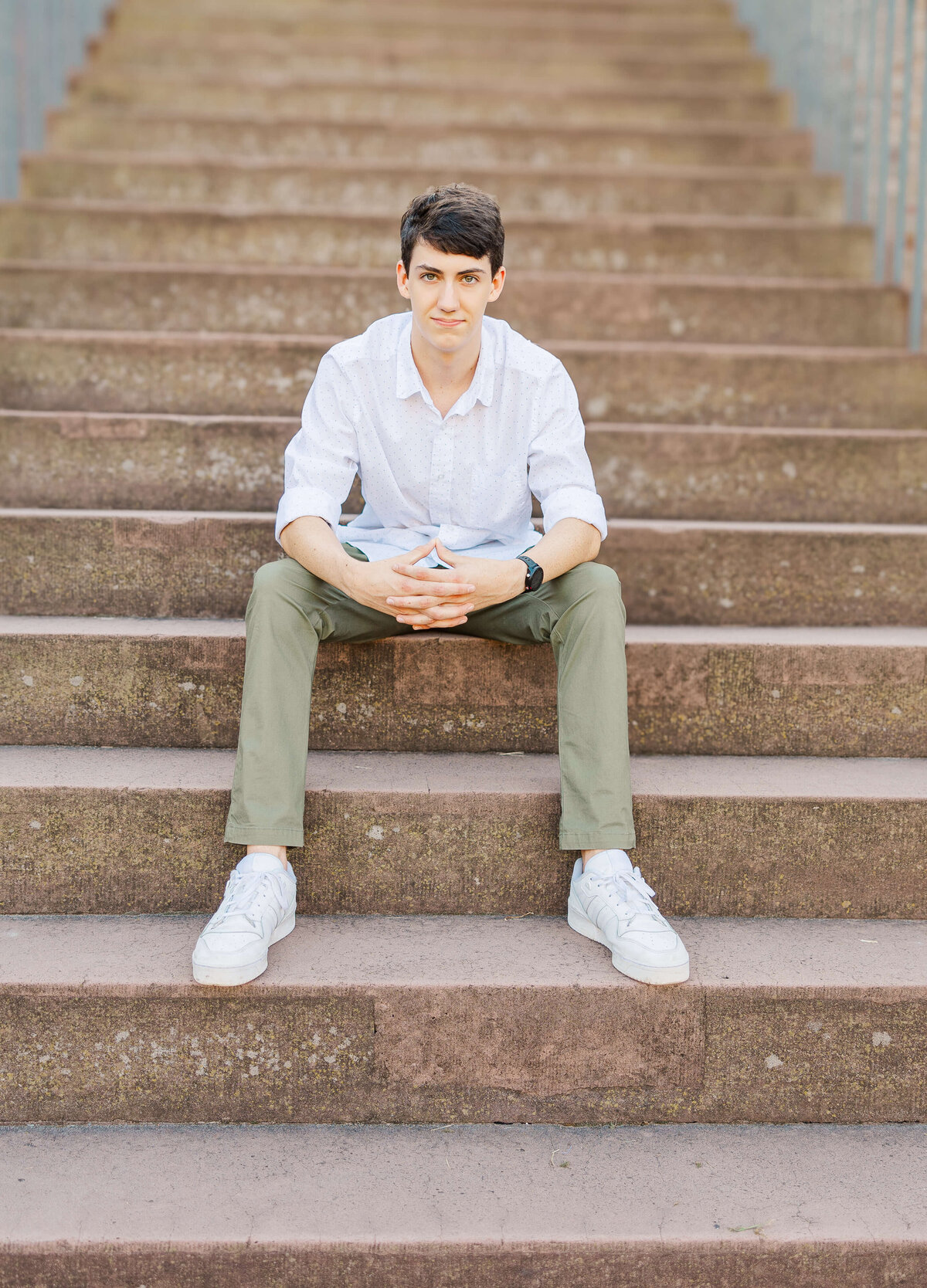 A person with short dark hair sits on wide stone steps, wearing a white shirt, khaki pants, and white sneakers. Their hands are clasped together as they look toward the camera of a senior photographer in Fayetteville, NC. The background is a set of stone stairs.
