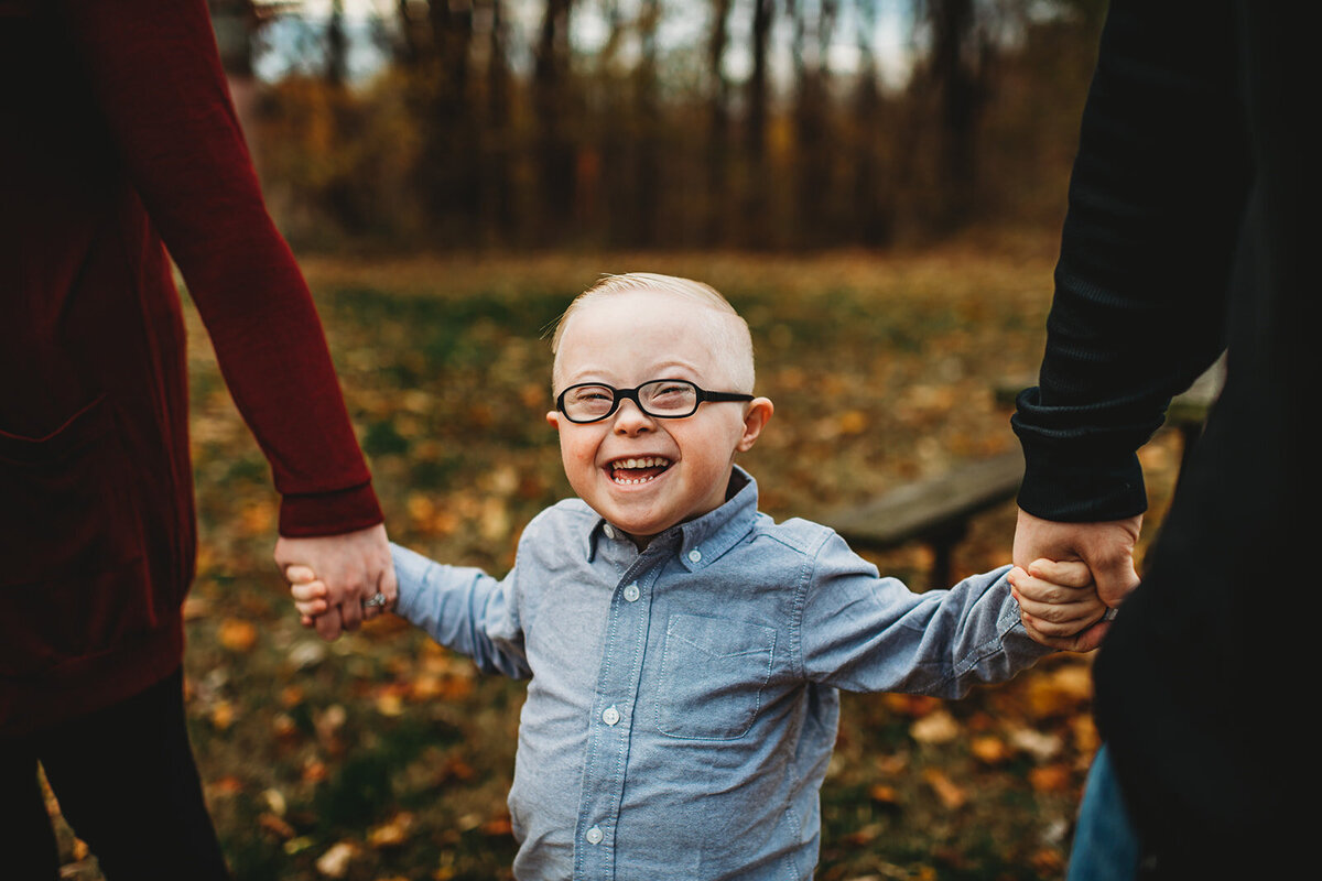 Family photographers Maryland captures little toddler child smiling as he holds hand with his parents in a park for fall family pictures