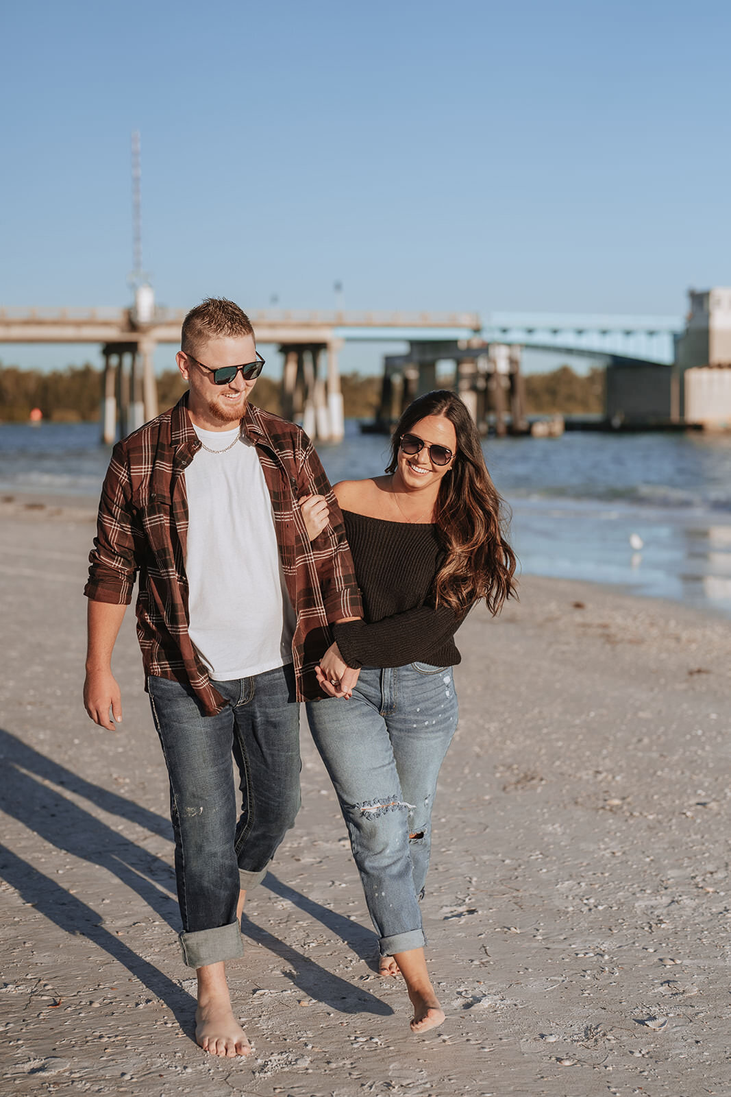 couple holding hands and walking on the beach with sunglasses on during engagement photos