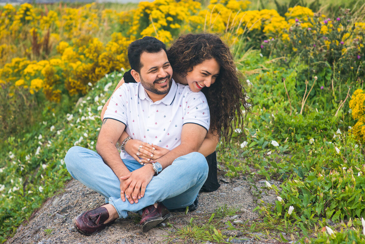 asian couple sitting on a rock surrounded by yellow flowers lokking away from camera laughing