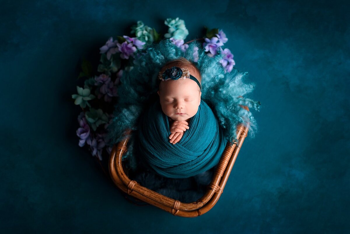 Newborn girl in jade teal potato wrap with lavendar flowers in bamboo basket.
