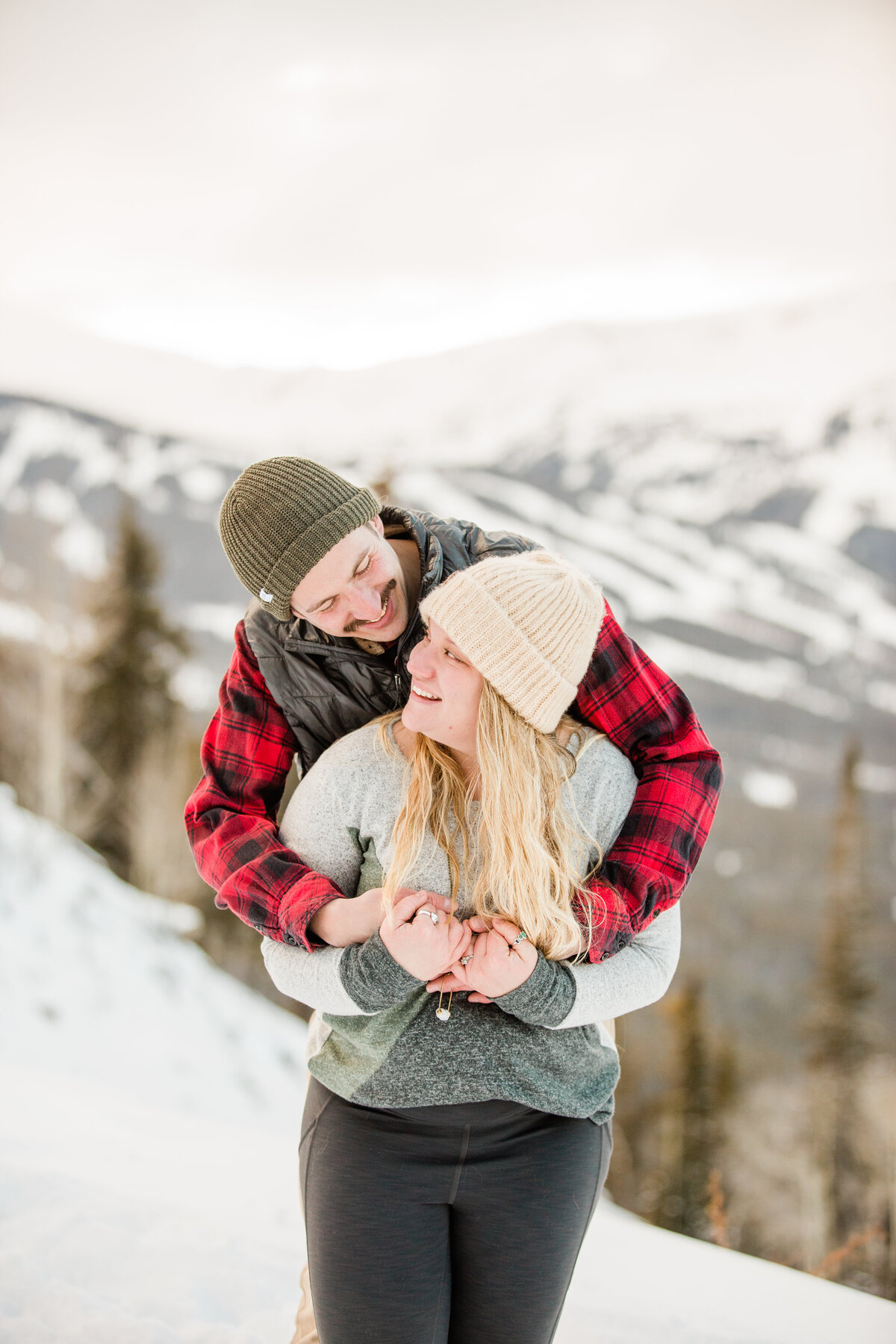 A man is embracing is fiance from behind. They are looking at one another smiling, with Breckenridge Mountain behind them