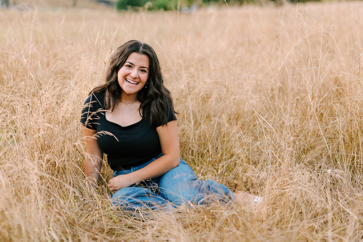 A teenage girl in jeans and a black tshirtsits in a field smiling