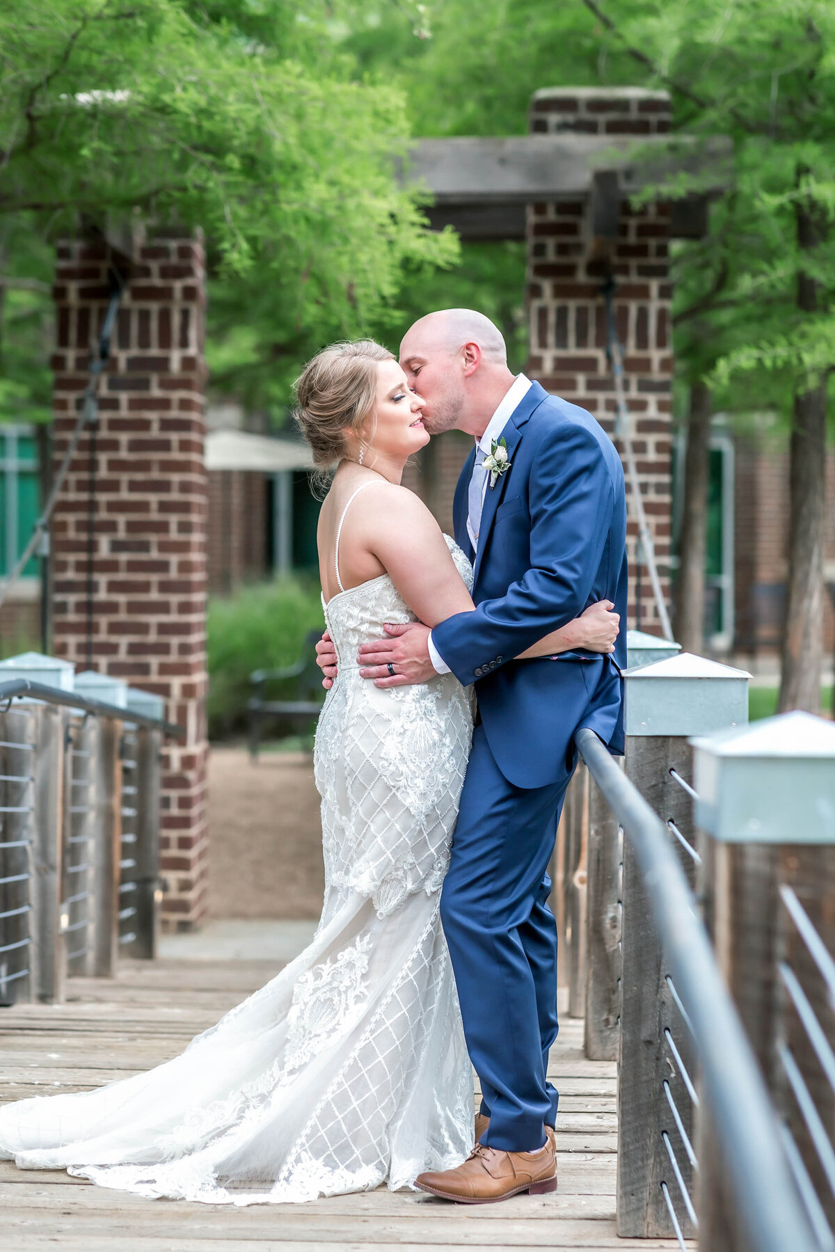 Bride is wearing a lace spaghetti strapped wedding dress and groom is wearing a blue suit.  Couple are standing on the bridge at the Willows Event Center in Lubbock Texas.  Groom is kissing bride on the cheek as they are facing each other.
