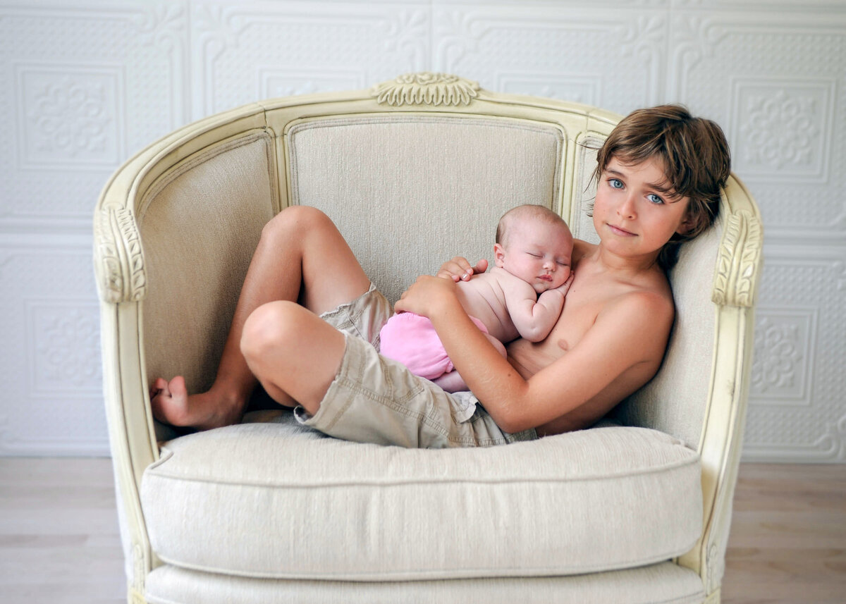 A young brown haired boy wearing khaki shorts and looking at the camera while holding his baby sitster in a large  cream colored chair.