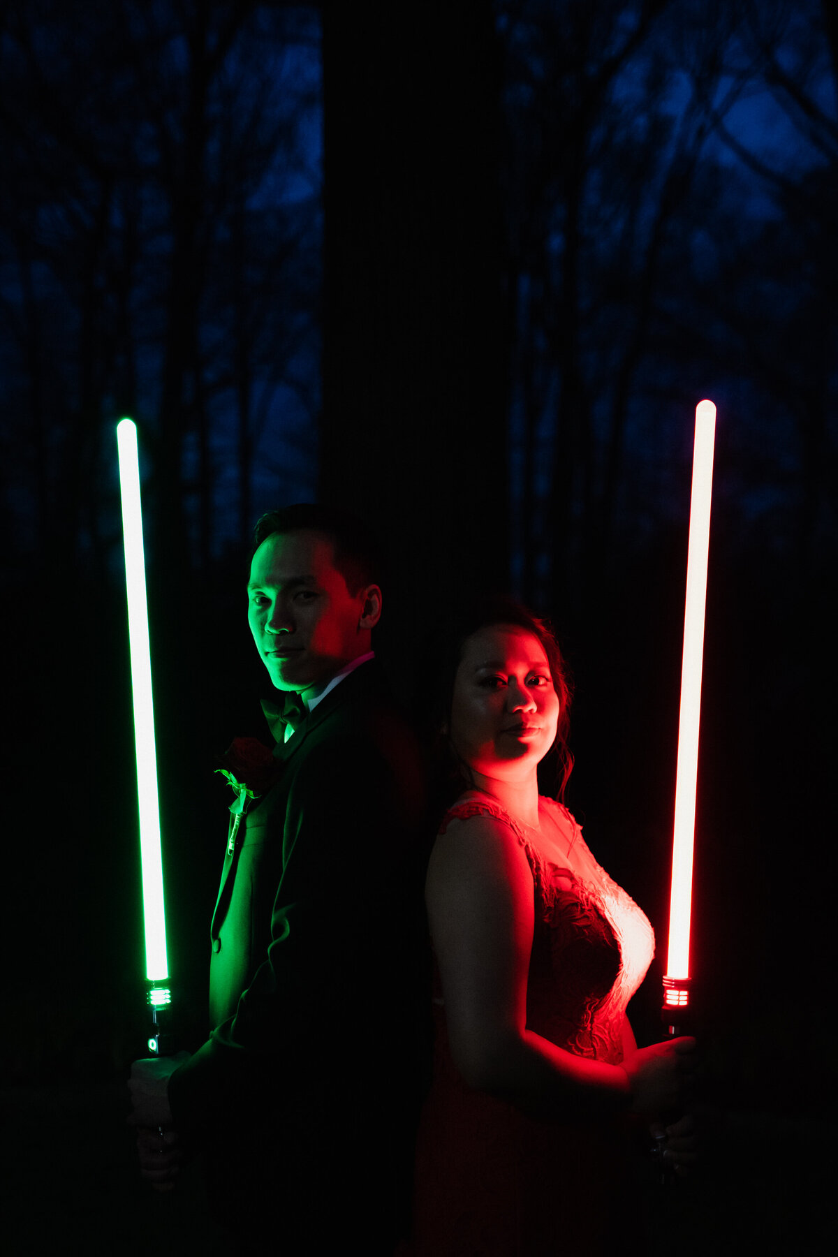 A couple standing in a dark forest holding lightsabers in front of them.