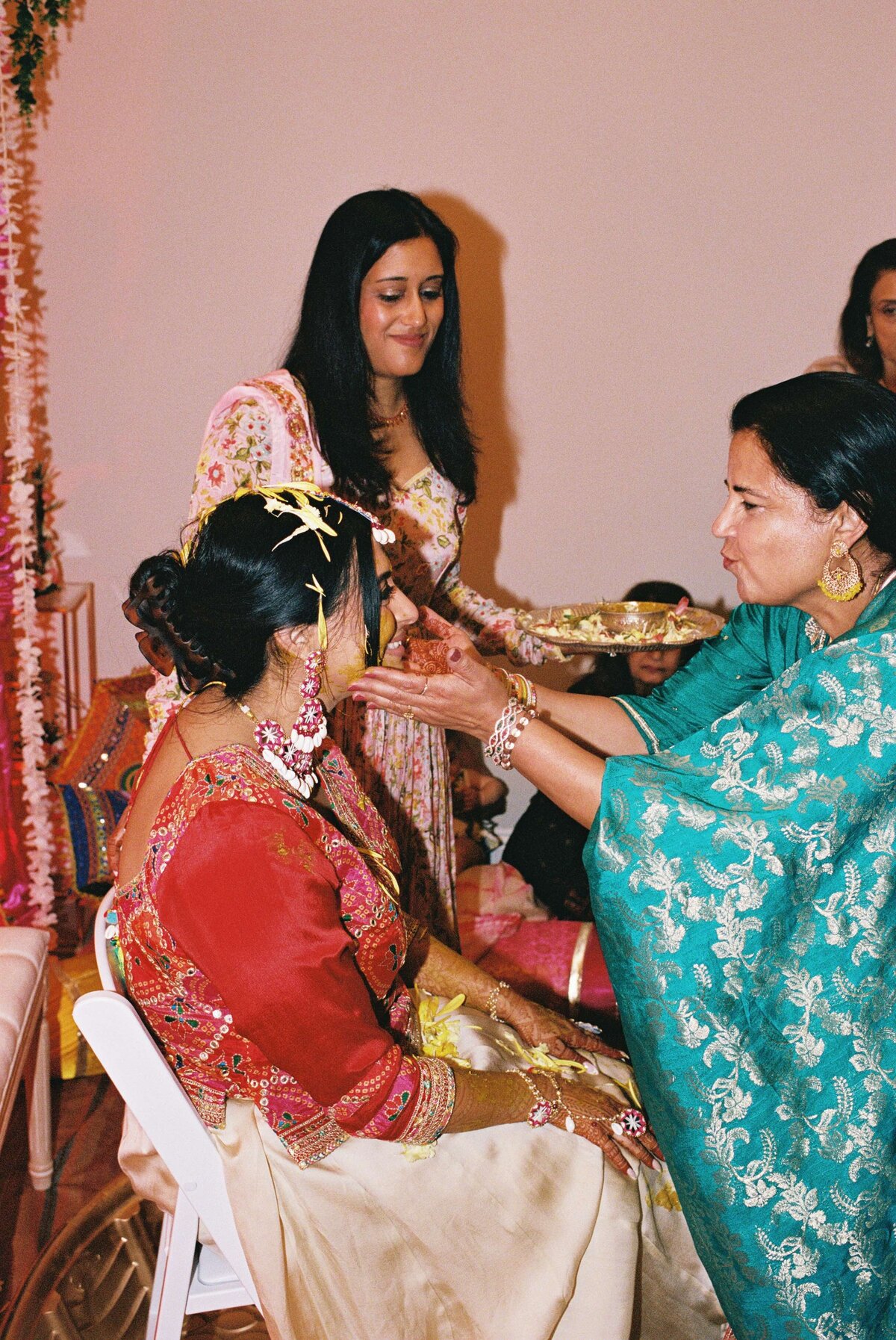 A woman in a red outfit sits on a chair, receiving a traditional blessing from another woman in blue. A third woman in floral attire stands nearby, smiling. The atmosphere is warm and celebratory.