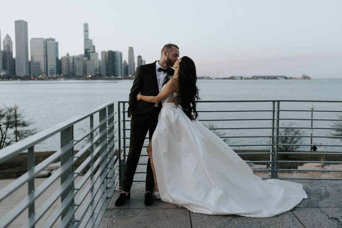 Husband and wife kiss on wedding day overlooking lake Michigan and the skyline.