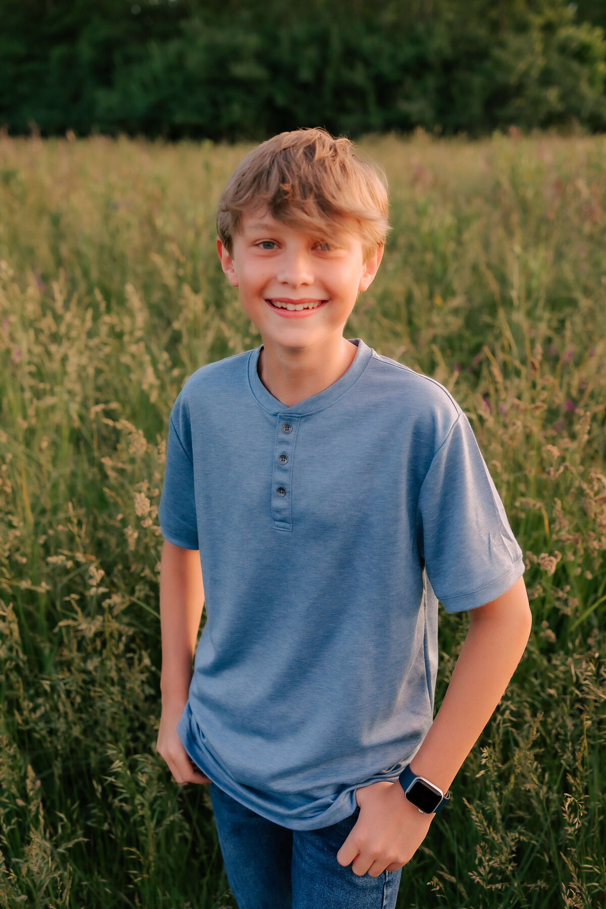 A handsome young man stands in the field surrounded by tall grass at sunset.