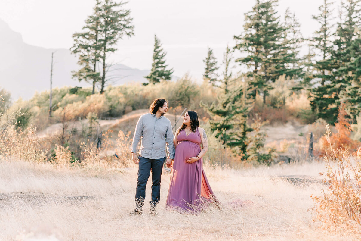 maternity portrait outdoors in the columbia river gorge . mom is wearing pink dress and holding husbands hand  while they look at each other