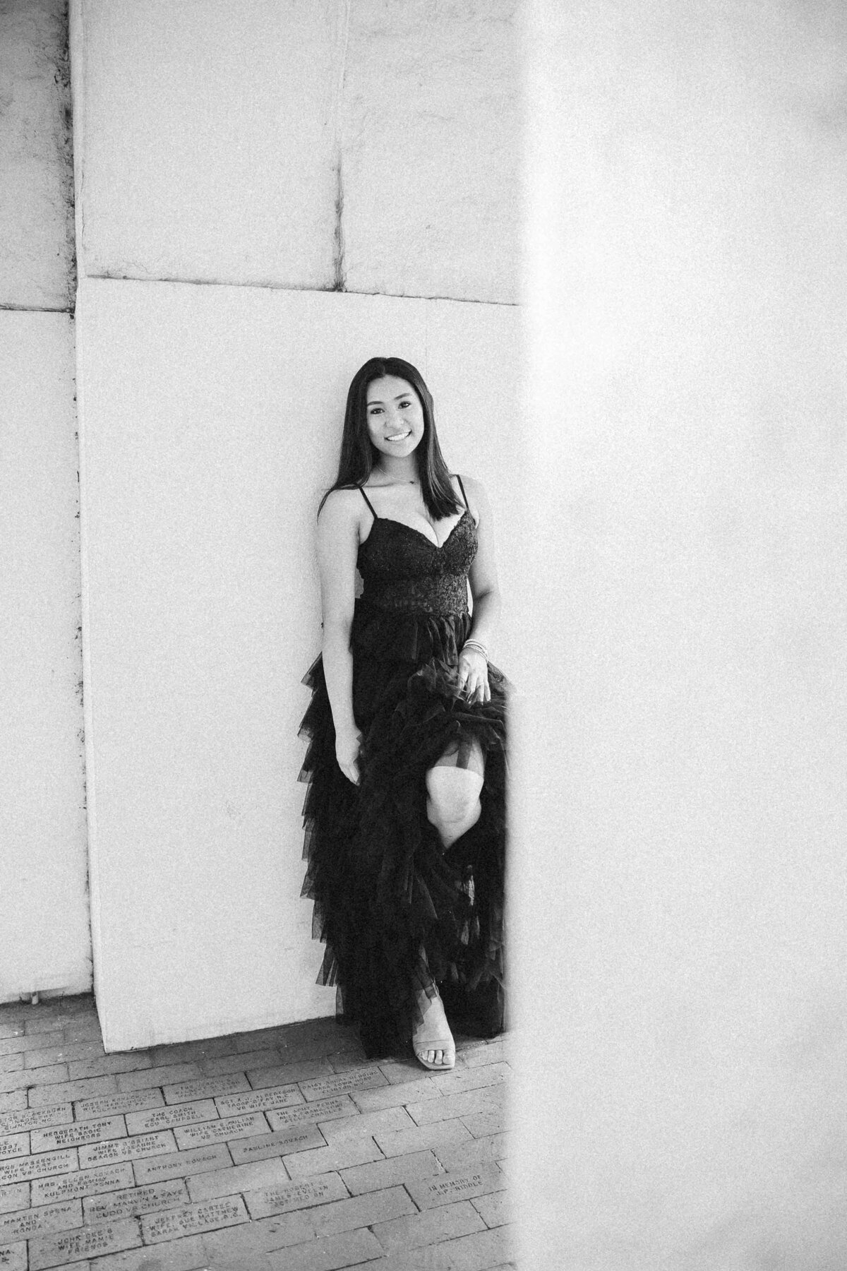 A person in a flowing black dress leans against a white pillar under an archway on a brick walkway, captured expertly by a senior photographer in Fayetteville, NC. The background is softly blurred, highlighting bright outdoor light and a green bush.