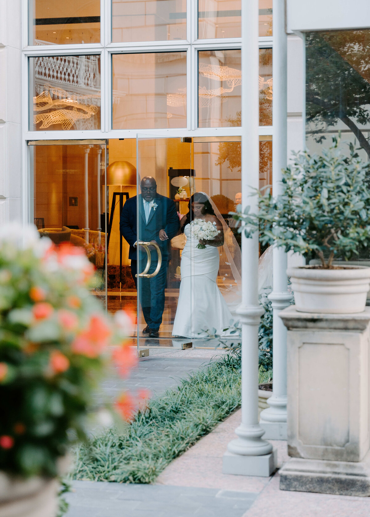 Bride in white gown and groom in dark suit stand behind glass doors, framed by vibrant flowers and green plants, creating a serene and joyful scene.