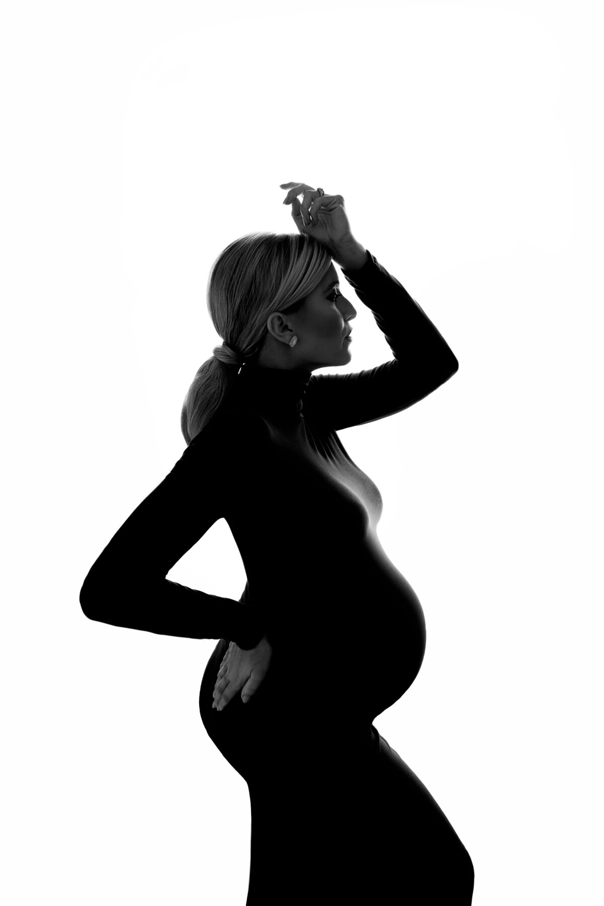 Woman poses for fine art maternity photos with Main Line's best maternity photographer Katie Marshall. Black and white image. Woman is standing side profile in a long-sleeved body con dress.  One hand is on the small of her back, the other hand has her wrist resting on her forehead. Her eyes are closed.