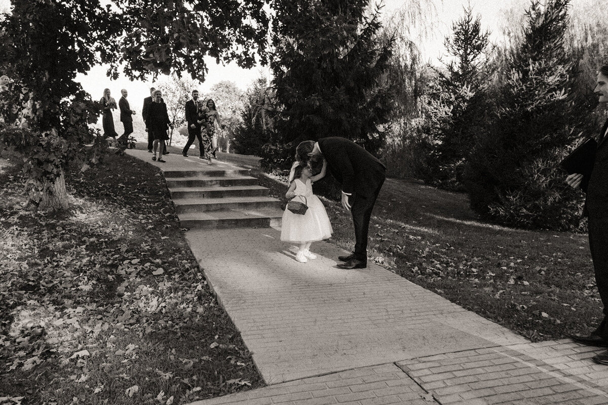 A black and white photo of a man bending down to kiss a child in a fluffy dress on a garden pathway at a park, with people walking in the background.