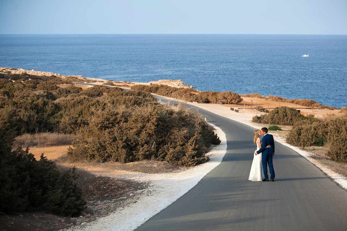 Bride & Groom kssing from a distance on a deserted road with the sea in the background