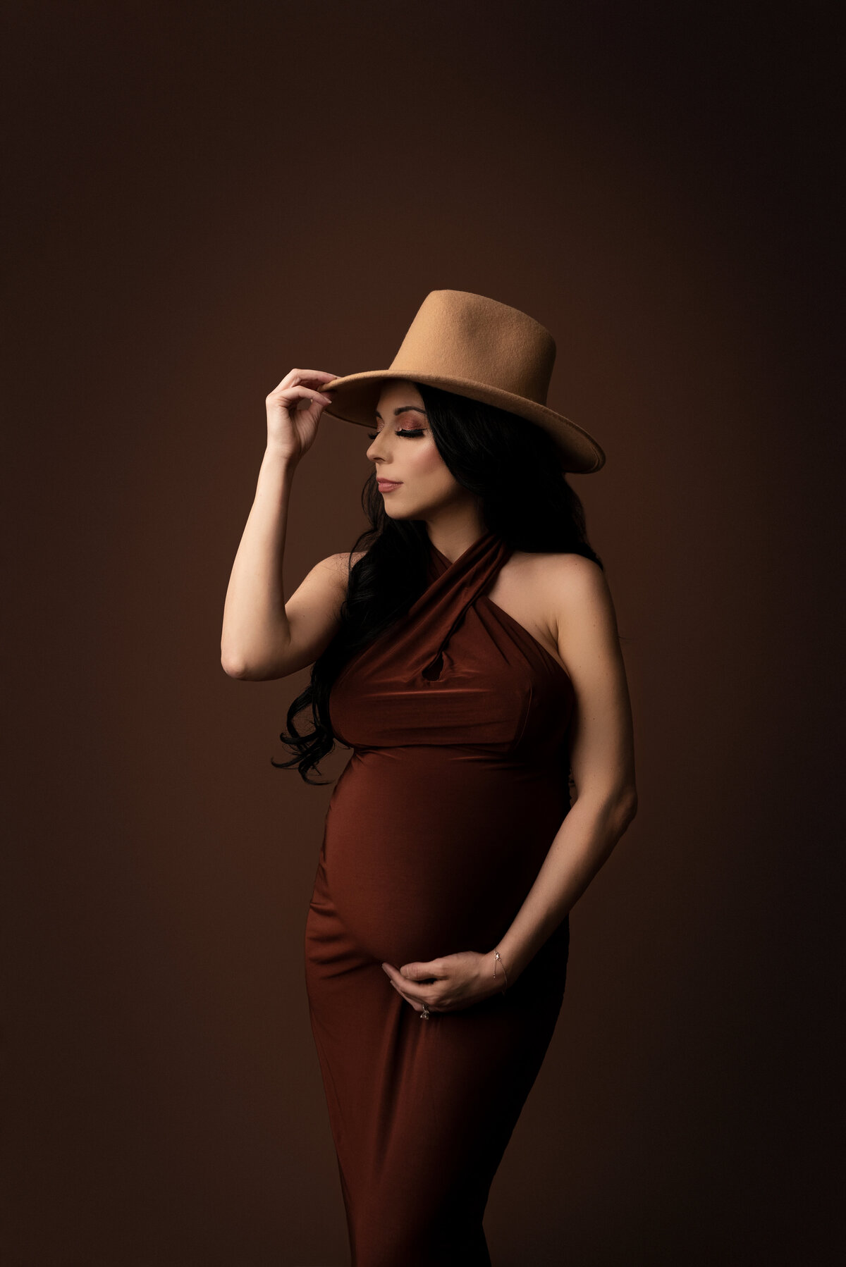 Captured by Katie Marshall, renowned as the best Philadelphia Main Line maternity photographer, this stunning black and white maternity photo features a woman standing against a rich brown backdrop. She is elegantly attired in a burgundy-brown halter maternity gown and a stylish tan wide-brim hat, creating a captivating contrast. Slightly angled away from the camera, one hand cradles her baby bump, while the other touches the brim of her hat. Her head is turned away from the camera, showcasing her graceful side profile, resulting in a powerful and emotive maternity portrait.