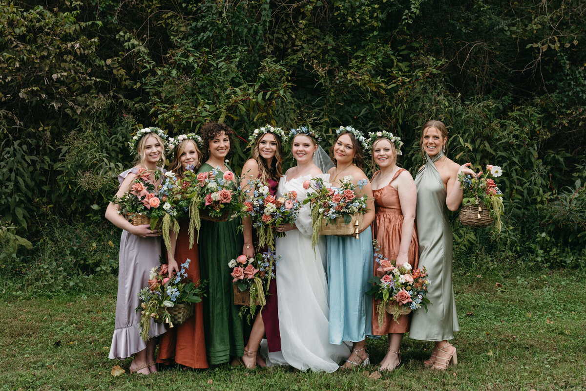 Bridal party group photo featuring bridesmaids in mismatched jewel-tone dresses holding lush garden-style bouquets with roses, delphinium, dahlias, and greenery. Bride stands center with a romantic floral crown and meadow-inspired bouquet. Captured against a woodland backdrop in Northwest Arkansas.