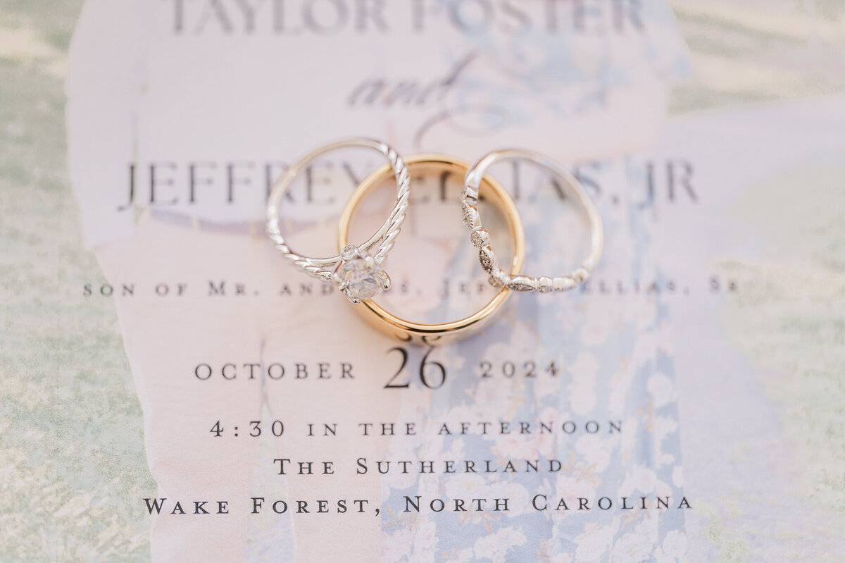 An engagement ring next to a gold wedding band on top of wedding invitations by Raleigh wedding photographer, JoLynn Photography