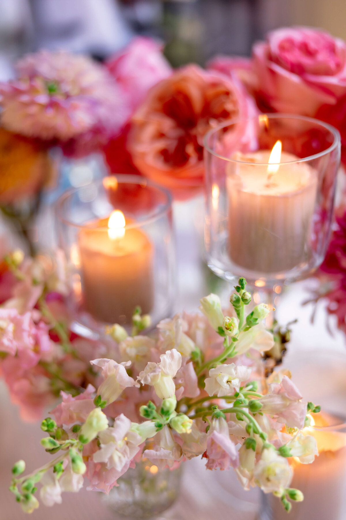 Candles glow in glass holders surrounded by an arrangement of pink, peach, and orange flowers. Bright and delicate blossoms, including roses and snapdragons, create a romantic, serene ambiance.