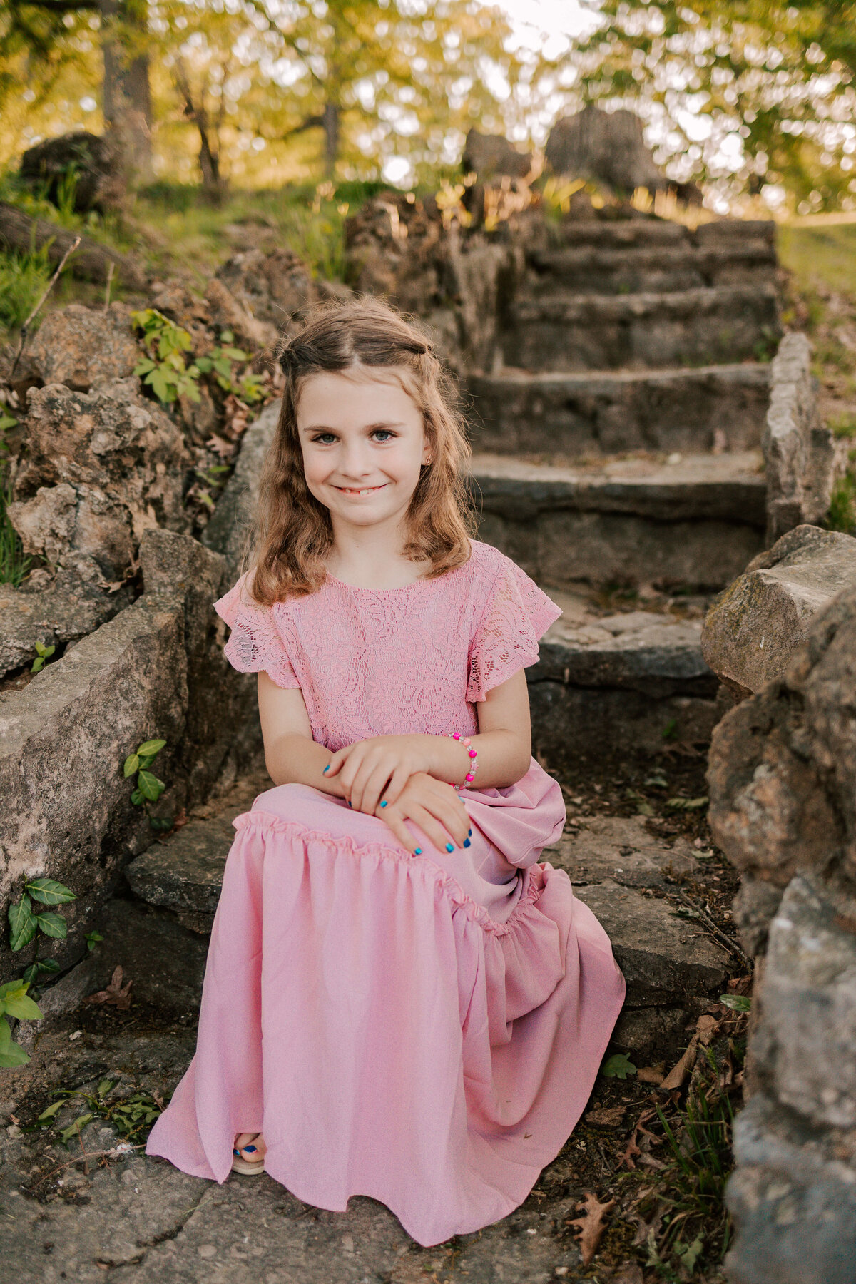 A beautiful girl wearing a long pink dress is sitting on stone steps in a St. Louis park at sunset.