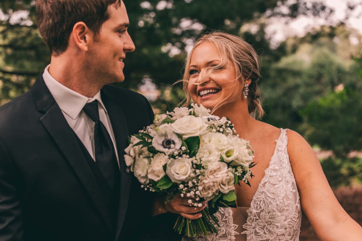 A wedding couple smiling and looking at each other, the bride holding a bouquet of white flowers, outdoors with greenery in the background, possibly planned by a wedding coordinator from Iowa.
