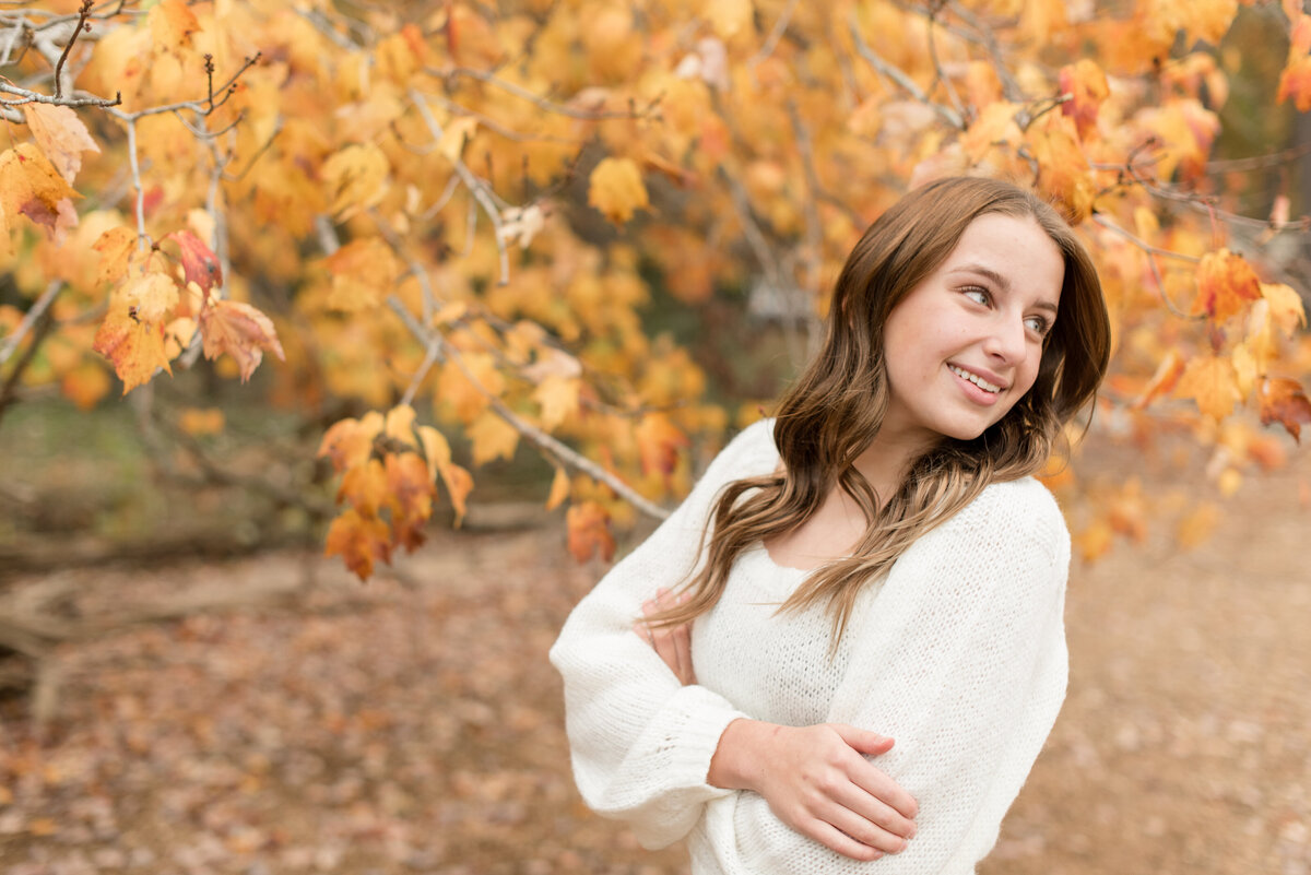 Senior girl leaning back and looking over left shoulder by bright yellow Fall tree in Lancaster, Pennsylvania.