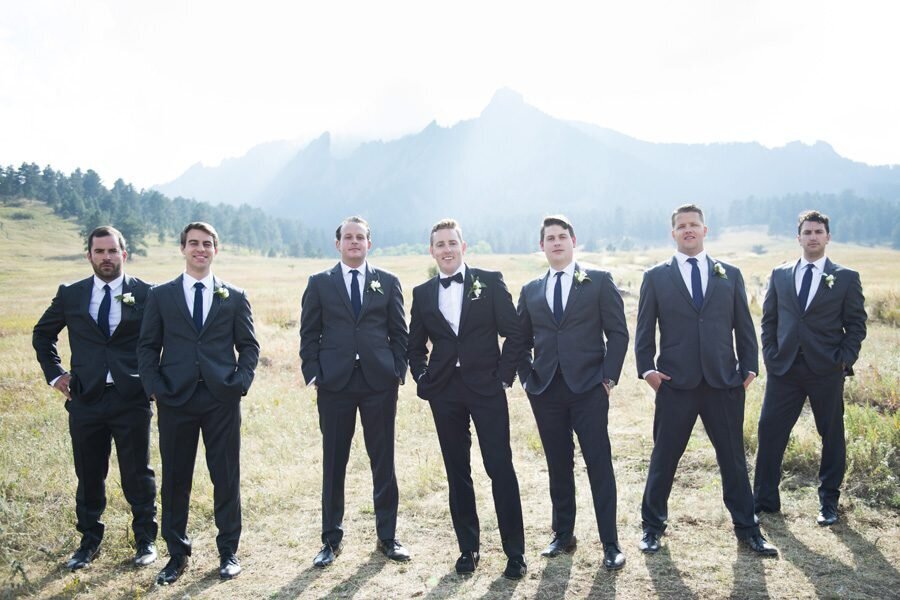 A groomsmen stands with his groomsmen in a field at The Manor House. All the men have their hands in their pockets