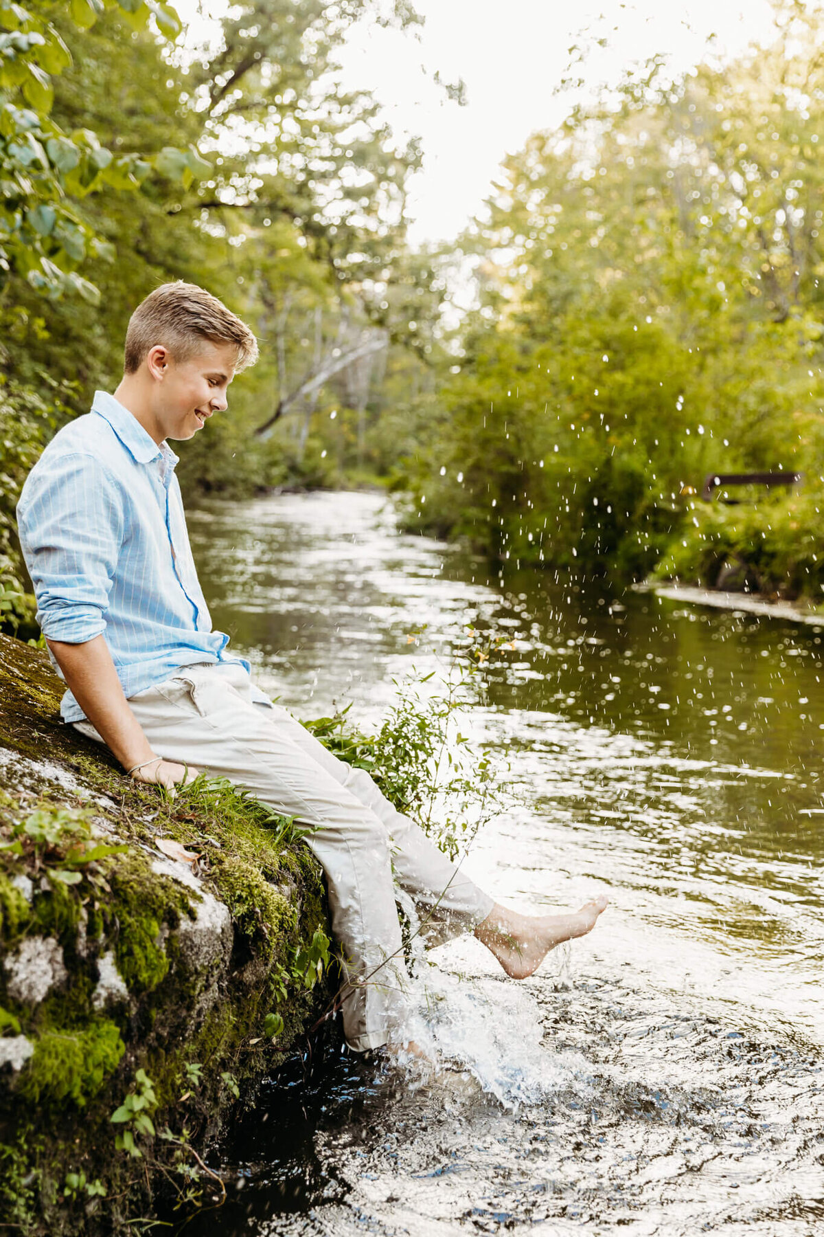 senior guy in a blue shirt and khakis sitting on the edge of a rock and splashing his feet in the water captured by Green Bay senior photographer Ashley Kalbus