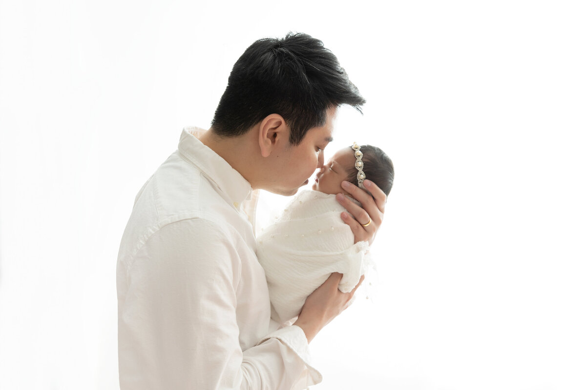 A new father leans in to kiss his sleeping newborn baby girl in his hands while standing in a studio