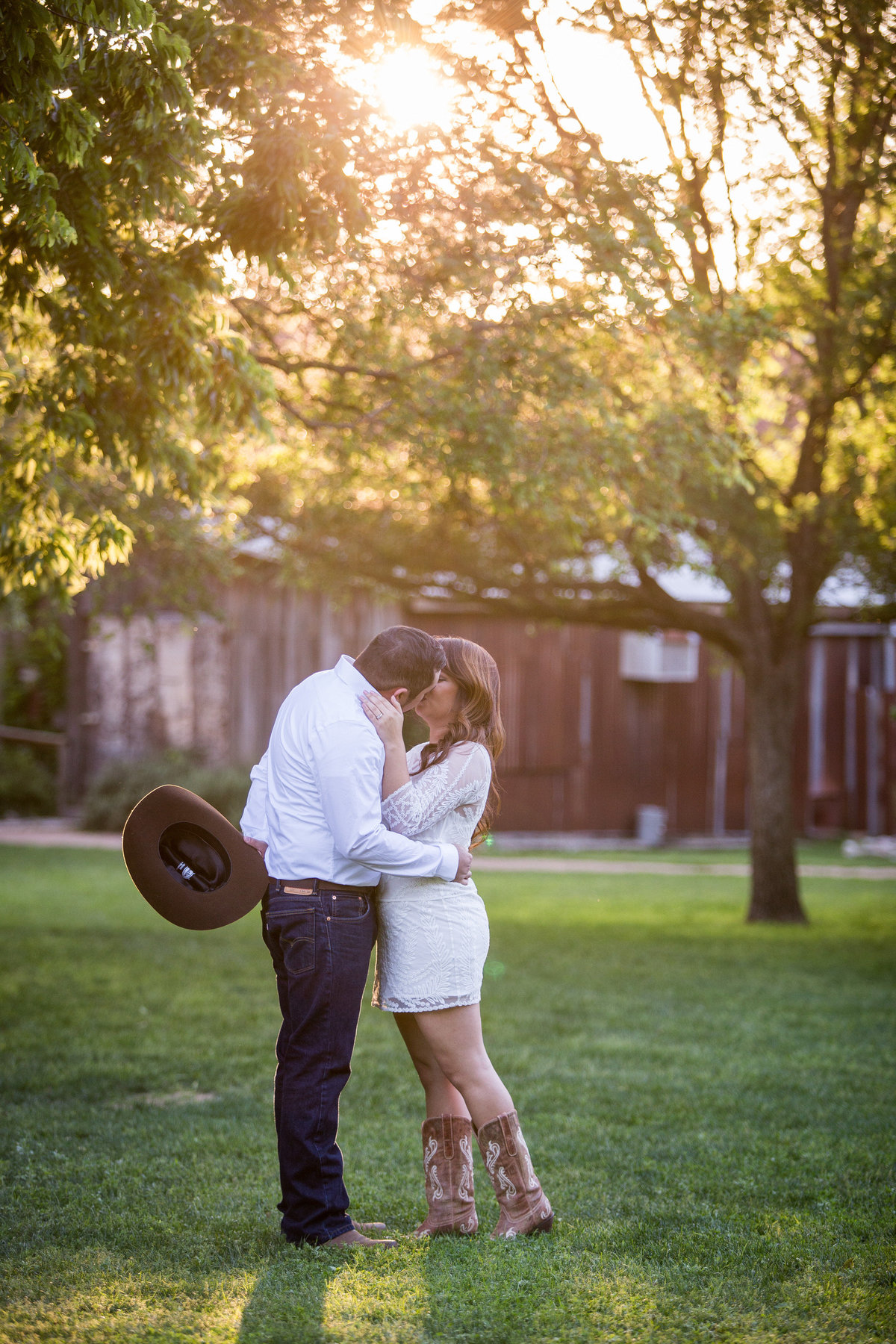 Engaged couple kissing in Gruene, Texas. Man is holding cowboy hat behind his back and sun is shining through trees.