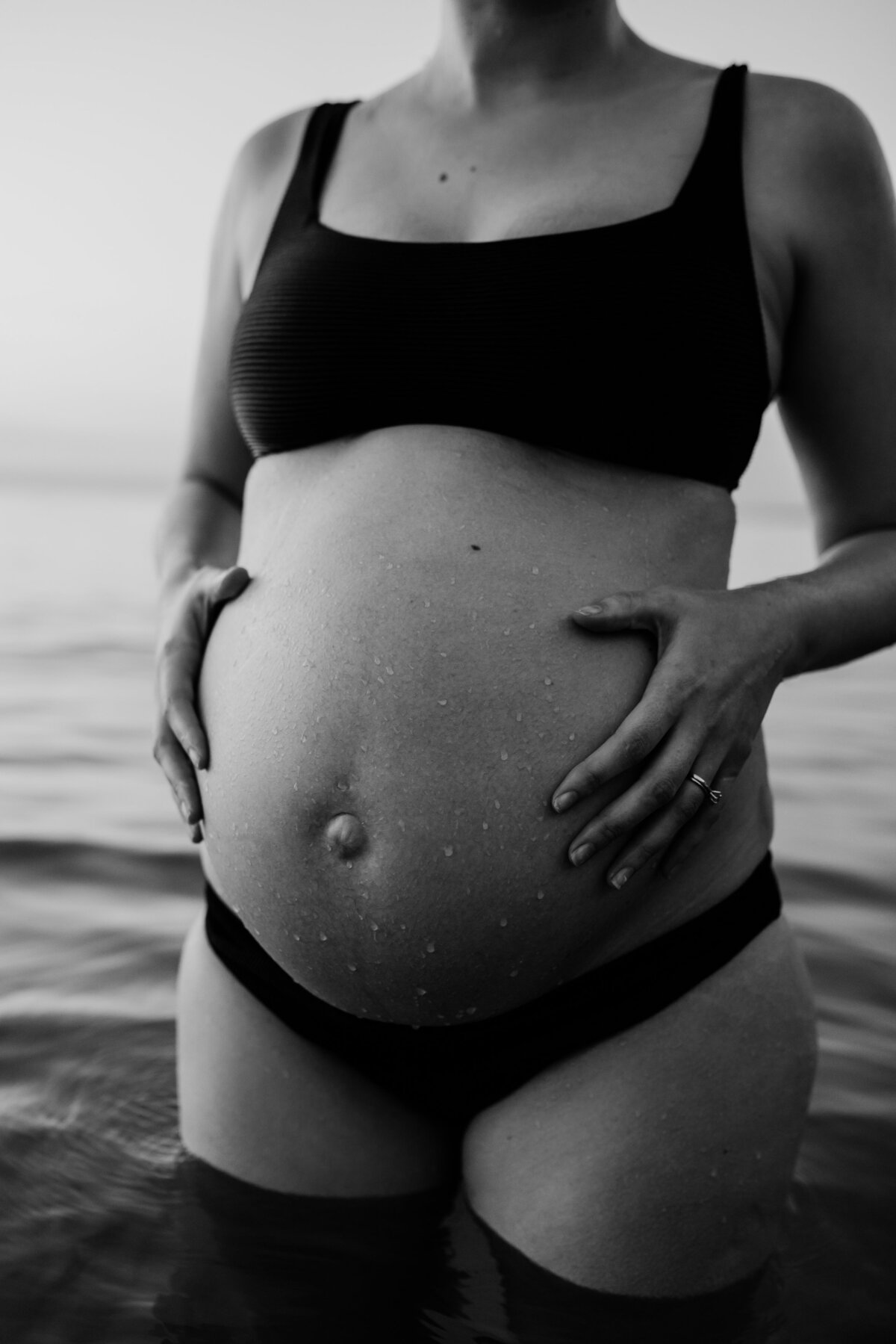 black and white image of pregnant woman wearing bikini, standing in water and holding belly.