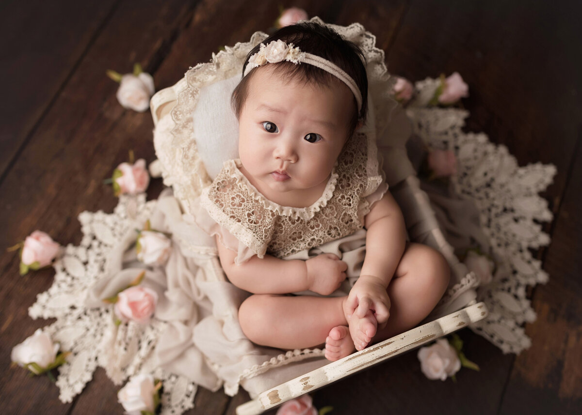 3-month baby girl milestone photoshoot in West Palm Beach and Jupiter, FL. Asian baby girl is in a lace romper curled up in a miniature vintage doll cradle. Baby is looking intently at the camera.