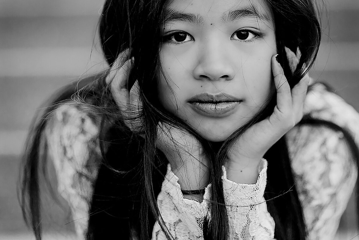 Close-up black and white photo of a girl with long , dark hair looks into the camera with her chin in her hands, cropped above her eyes.