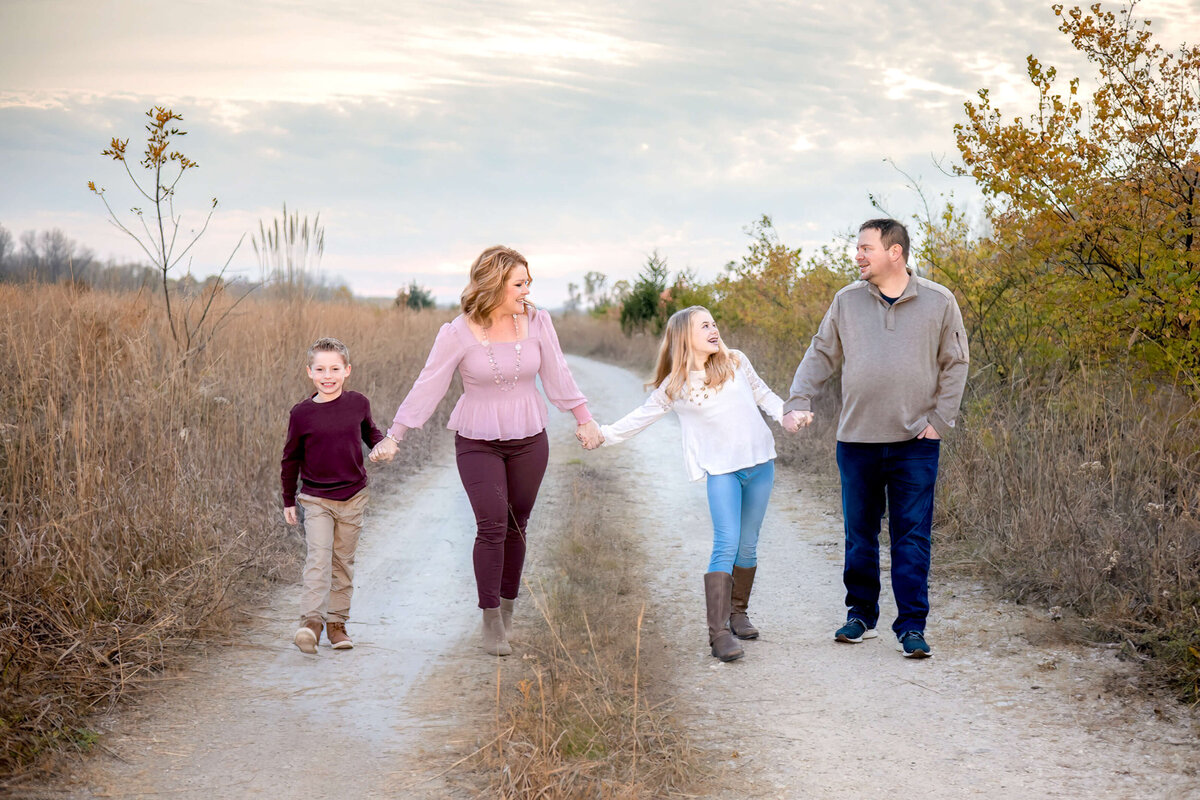 family of 4 waling hand in hand down a dirt path looking at each other and laughing and smiling