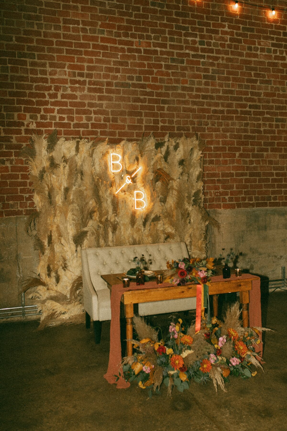 A wedding reception setup designed by a top wedding coordinator in Iowa, with a floral table, couch, and a backdrop featuring illuminated initials "b&amp;b" on a brick wall.