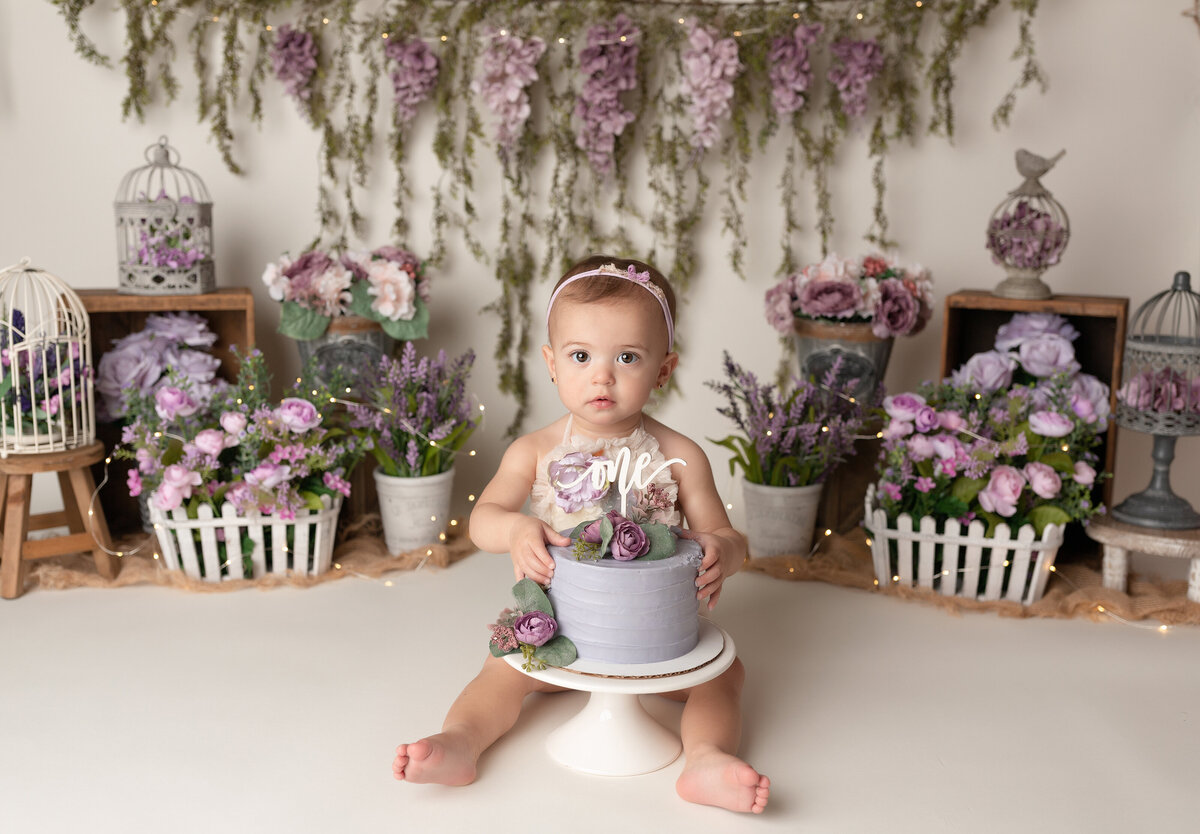 Elegant purple floral cake smash in West Palm Beach and Jupiter, FL photography studio. Baby girl is sitting behind a lavender cake decorated with real purple flowers. The baby is touching the cake looking at the camera. In the background there are baskets and buckets of purple and pink toned flowers and a wisteria garland draped across the backdrop.