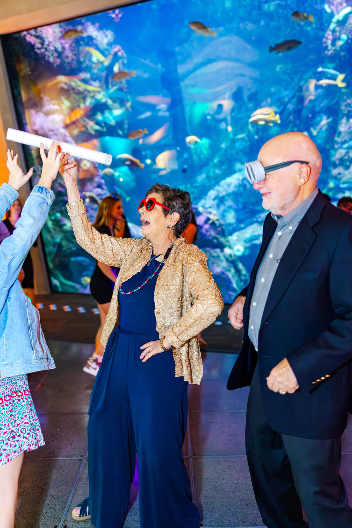 A woman in a blue suit and gold jacket dances with light tubes and big sunglasses at an aquaarium