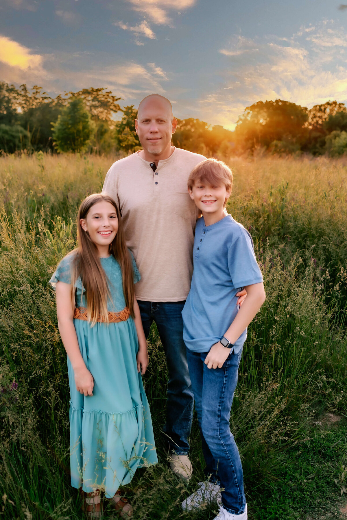 A father and his two kids pose for their family photo session in a field at sunset.