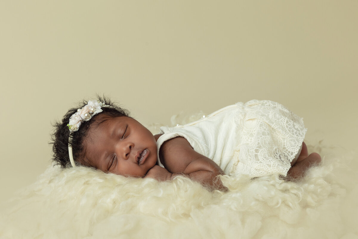 A newborn baby girl in a white lace dress sleeps on her stomach on a white blanket
