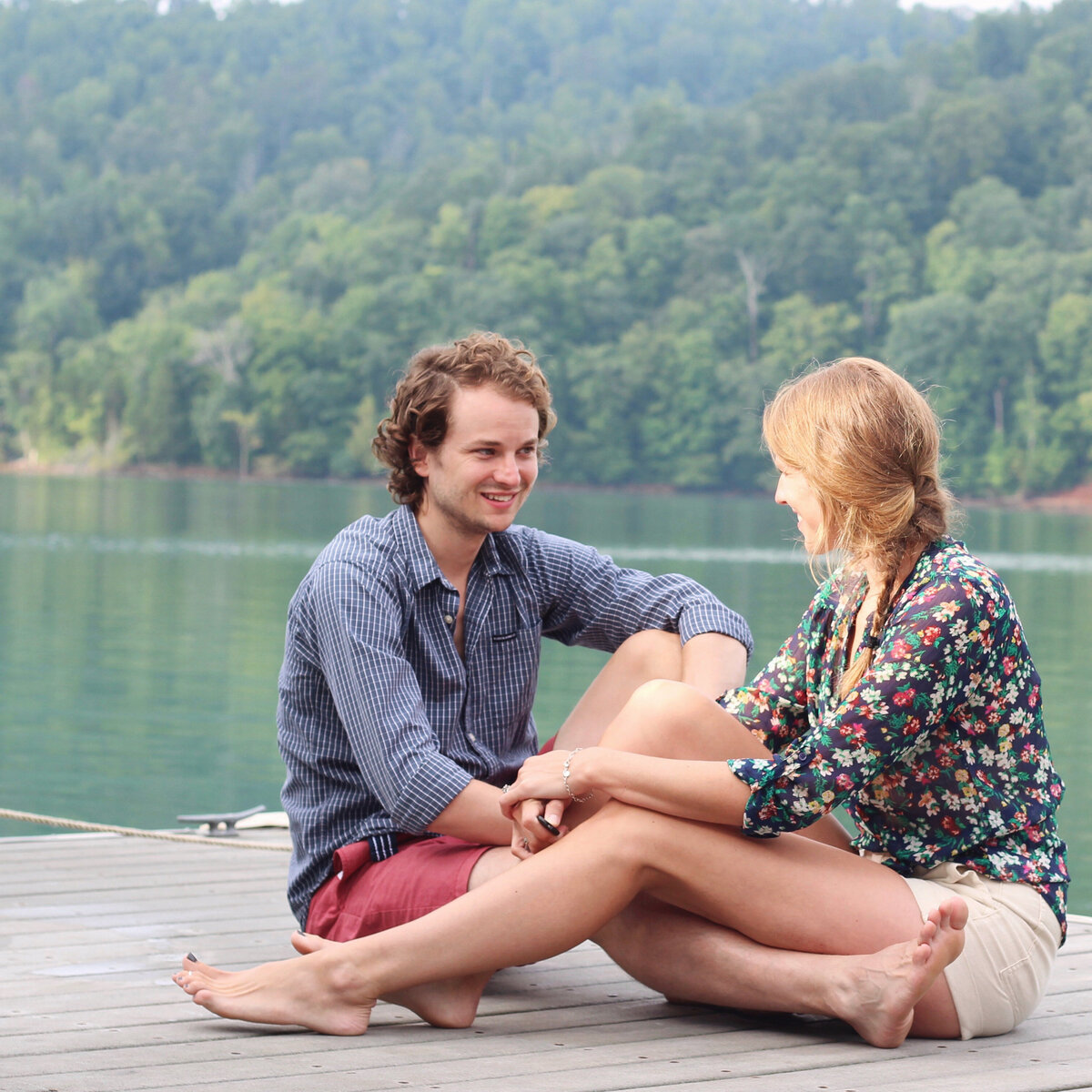 Couple sitting on lake dock smiling at one another with water and tree landscape behind them