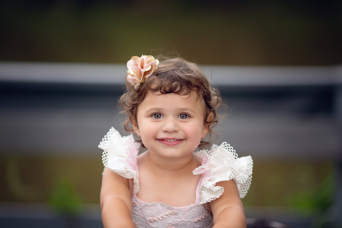 A toddler girl smiles big while wearing a pink flower in her hair to match her pink dress in a park