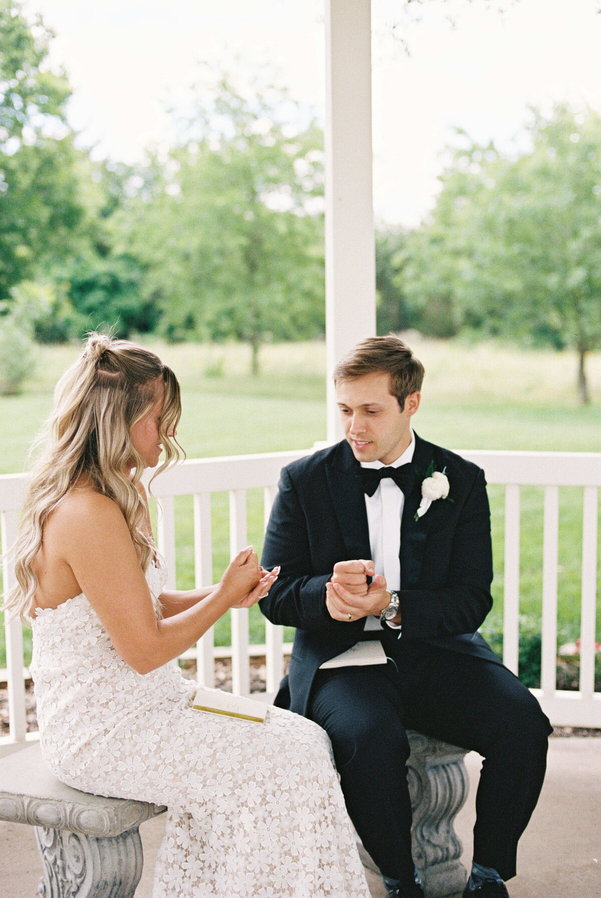 A bride in a white dress and a groom in a black tuxedo sit on a bench under a gazebo, exchanging notes. The background shows lush green trees, creating a serene and intimate atmosphere.