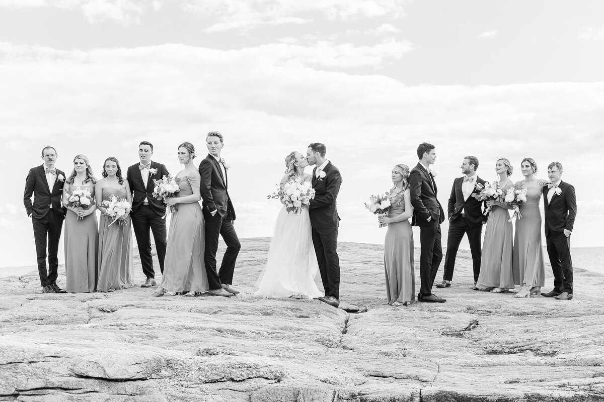 Bride and groom share a kiss on the rocks at the Madison Beach Hotel. The wedding party stands beside them looking in different directions. Captured by best New England wedding photographer Lia Rose Weddings.