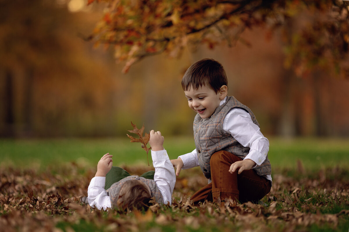 A young boy plays in a pile of leaves with his younger sibling in a park in fall