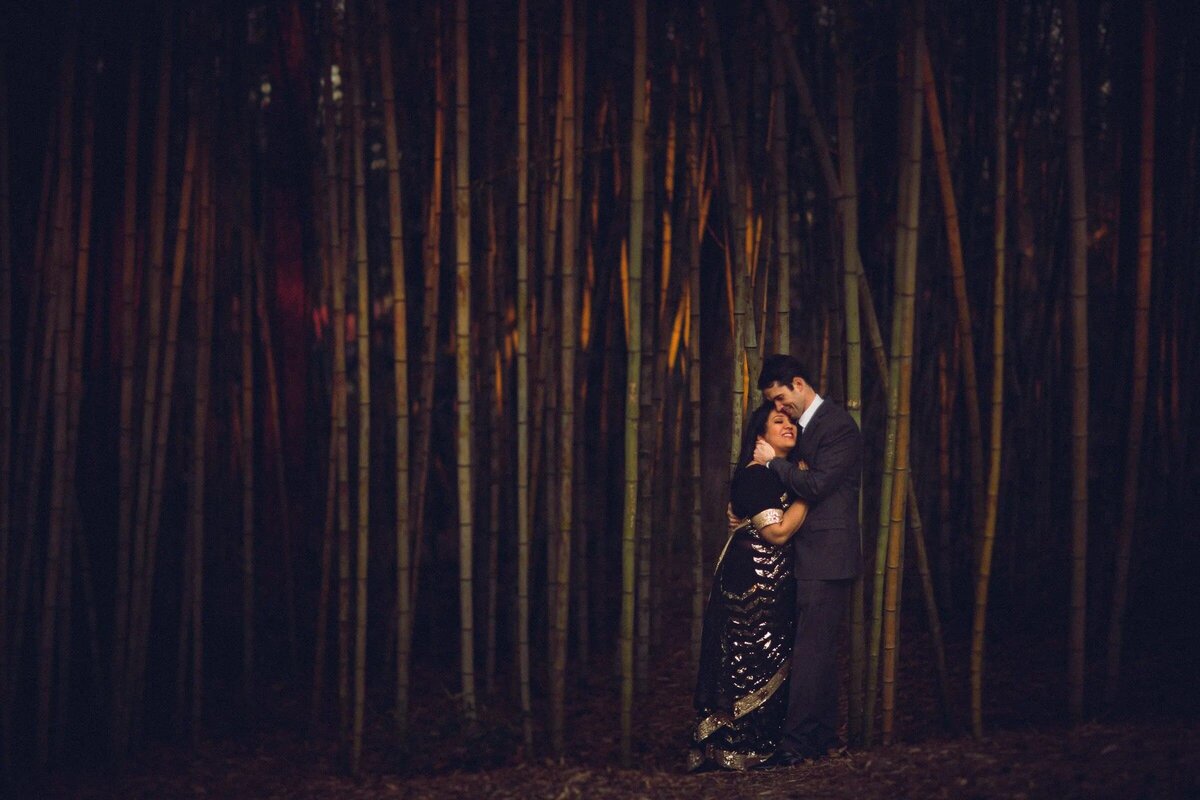 Intimate moment between a couple standing close in a dark bamboo forest, with a spotlight effect highlighting their embrace
