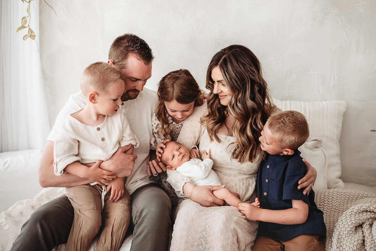 family of six with denver newborn photographer sitting on studio bed looking at baby brother
