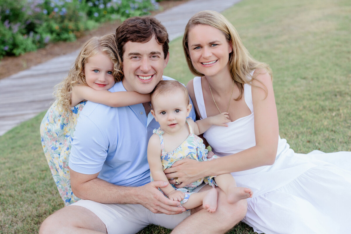 A small girl hanging on her dad back as he sits with his partner and a small baby